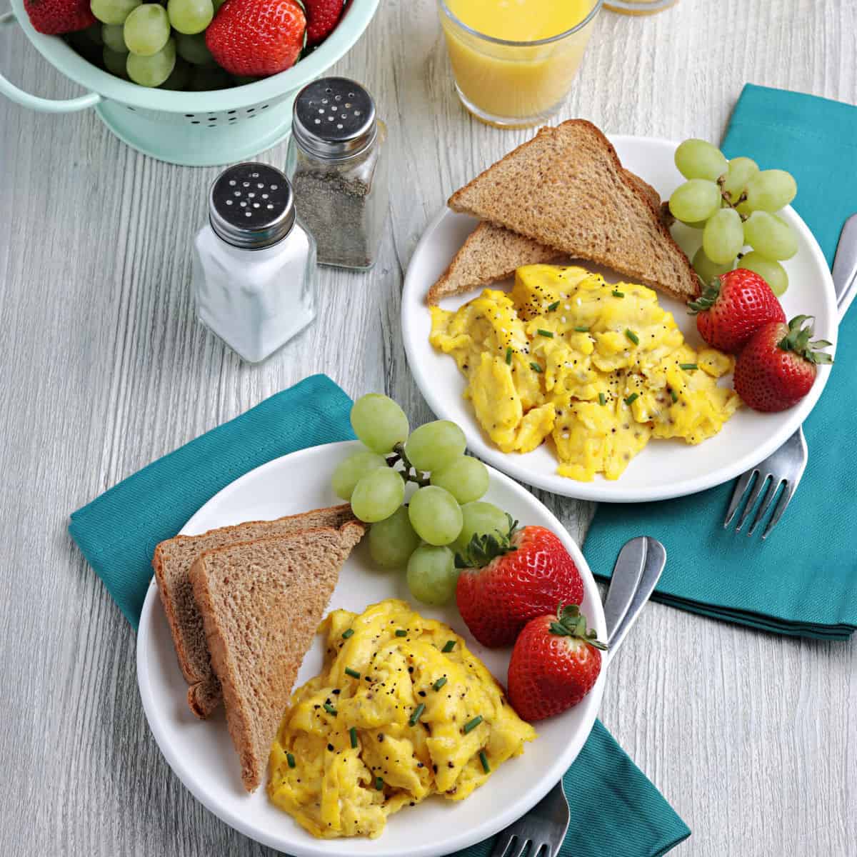 Two white plates of scrambled eggs with toast and fruit on the plate.