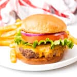 A cheeseburger hambuger with a bun on a white plate with lettuce, tomato and onion.