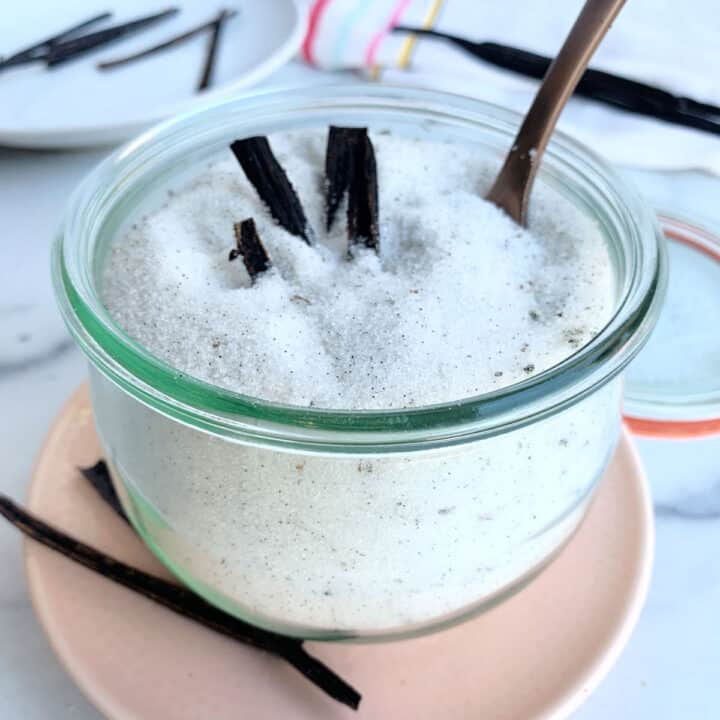 A glass jar of vanilla sugar, made with granulated sugar and vanilla bean pods, with a spoon inserted, sits on a pale pink plate.