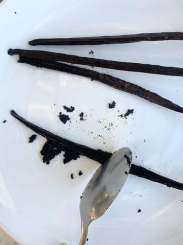 Scraping a vanilla bean on a white plate with a spoon.
