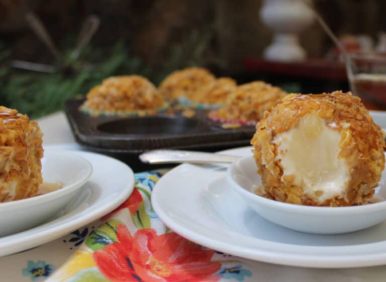 A scoop of fried ice cream coated in a crispy layer sits on a small white plate, with more servings in the background—one of those tempting no bake treats perfect for any dessert lover.
