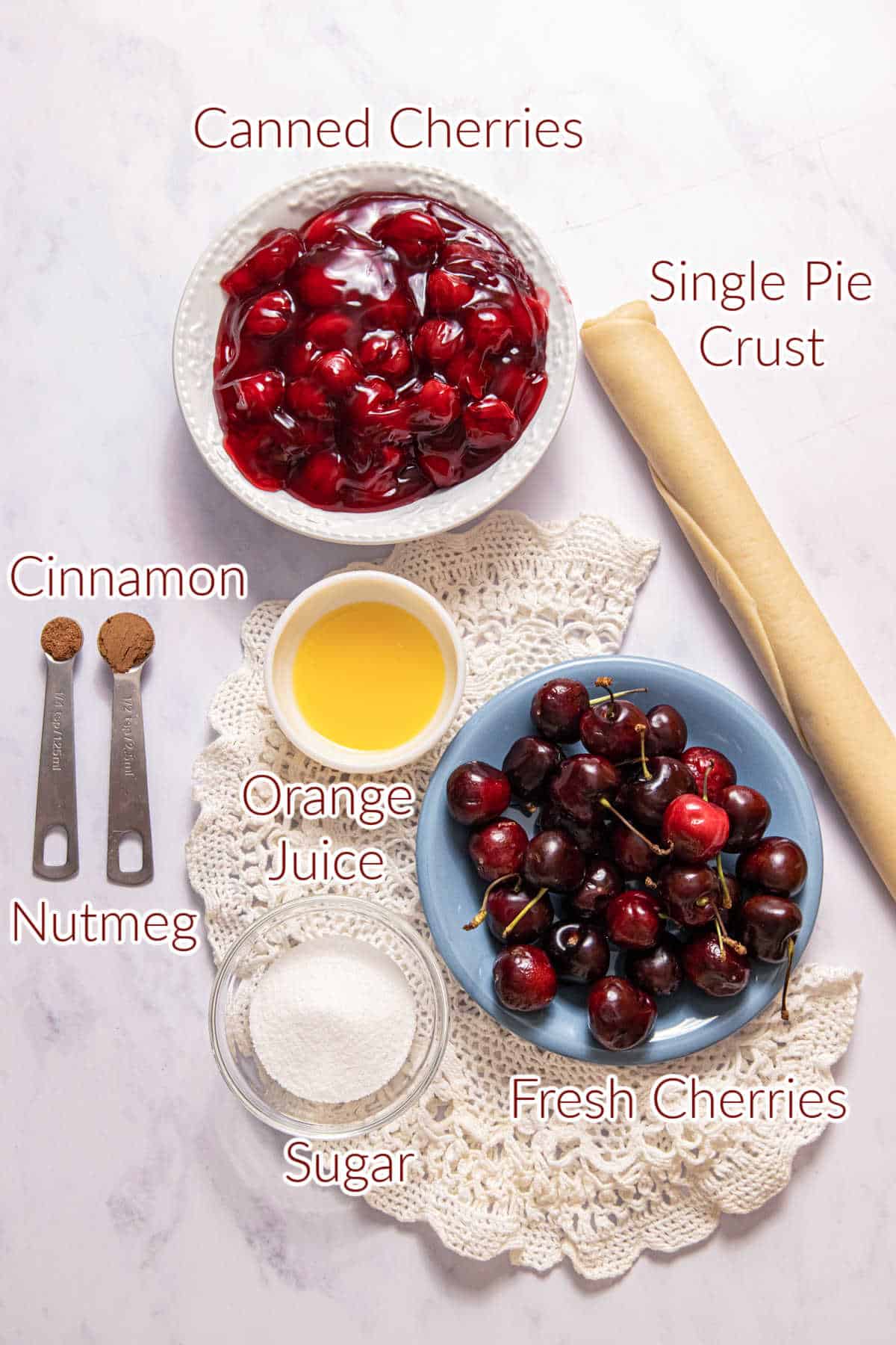 Ingredients for cherry pandowdy preparation laid out on a table, including canned cherries, fresh cherries, sugar, cinnamon, nutmeg, orange juice, and a pie crust.