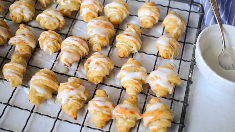 Freshly baked mini croissants with icing drizzle on a cooling rack.