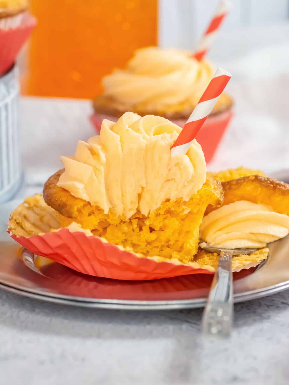 A partially eaten orange cupcake with swirled frosting and a red-striped straw sits on a plate, showcasing creative cake mix hacks; another cupcake and a drink appear in the background.