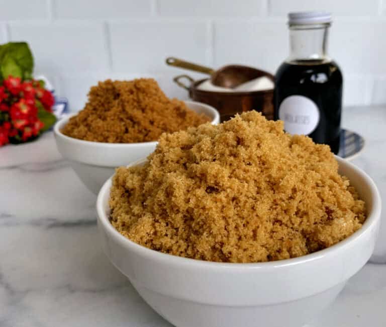 A close-up view of a bowl of brown sugar with more bowls in the background, a bottle of vanilla extract, and a decorative plant on a kitchen counter.