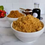Two bowls of brown sugar on a marble countertop, with a bottle of vanilla extract and wooden spoons in the background.