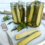 Two jars of homemade dill pickles with fresh dill on a plate, beside halved pickles and a fork, on a light background.