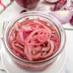 A jar of pickled red onions on a table surrounded by raw red onions.