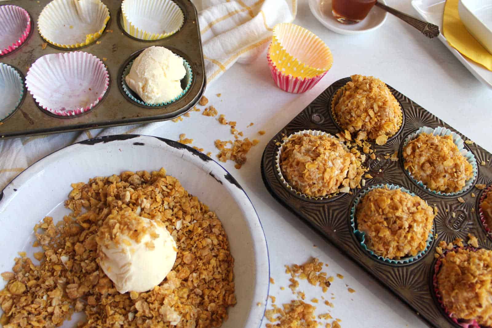 Two muffin trays are on a table, one filled with baked muffins and the other with cupcake liners and dough scoops. A bowl with crushed cornflakes and a scoop of dough is also visible.