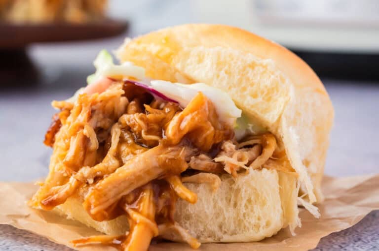 Close-up of a pulled pork sandwich with barbecue sauce and coleslaw, served on a white sandwich bun placed on a parchment paper.
