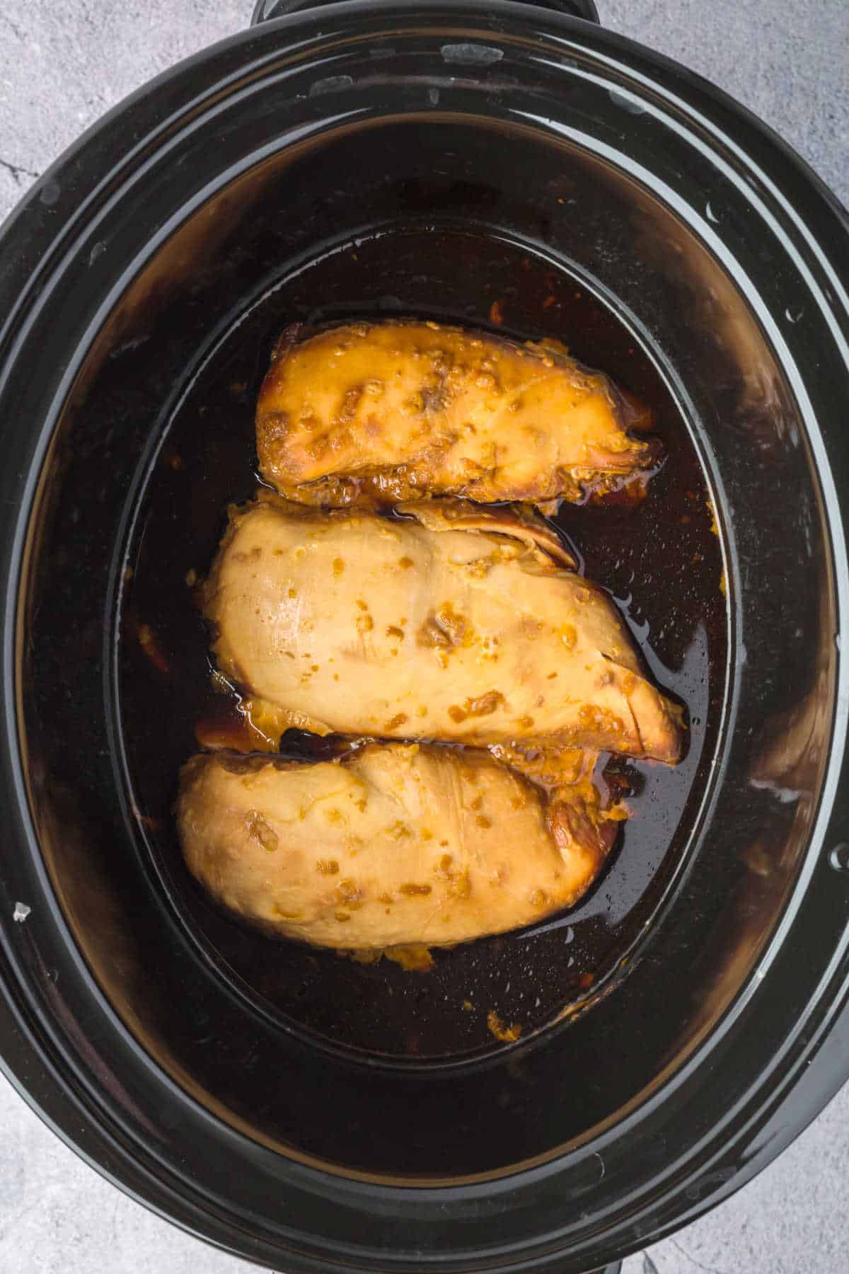 Three cooked chicken breasts in a dark brown sauce inside a black slow cooker.