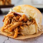A close-up of a pulled pork sandwich with coleslaw on a soft bun, placed on a piece of brown parchment paper.