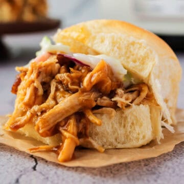 A close-up of a pulled pork sandwich with coleslaw on a soft bun, placed on a piece of brown parchment paper.