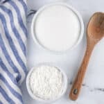 A bowl of sugar and a bowl of powdered sugar on a marble surface next to a wooden spoon and a blue striped cloth.