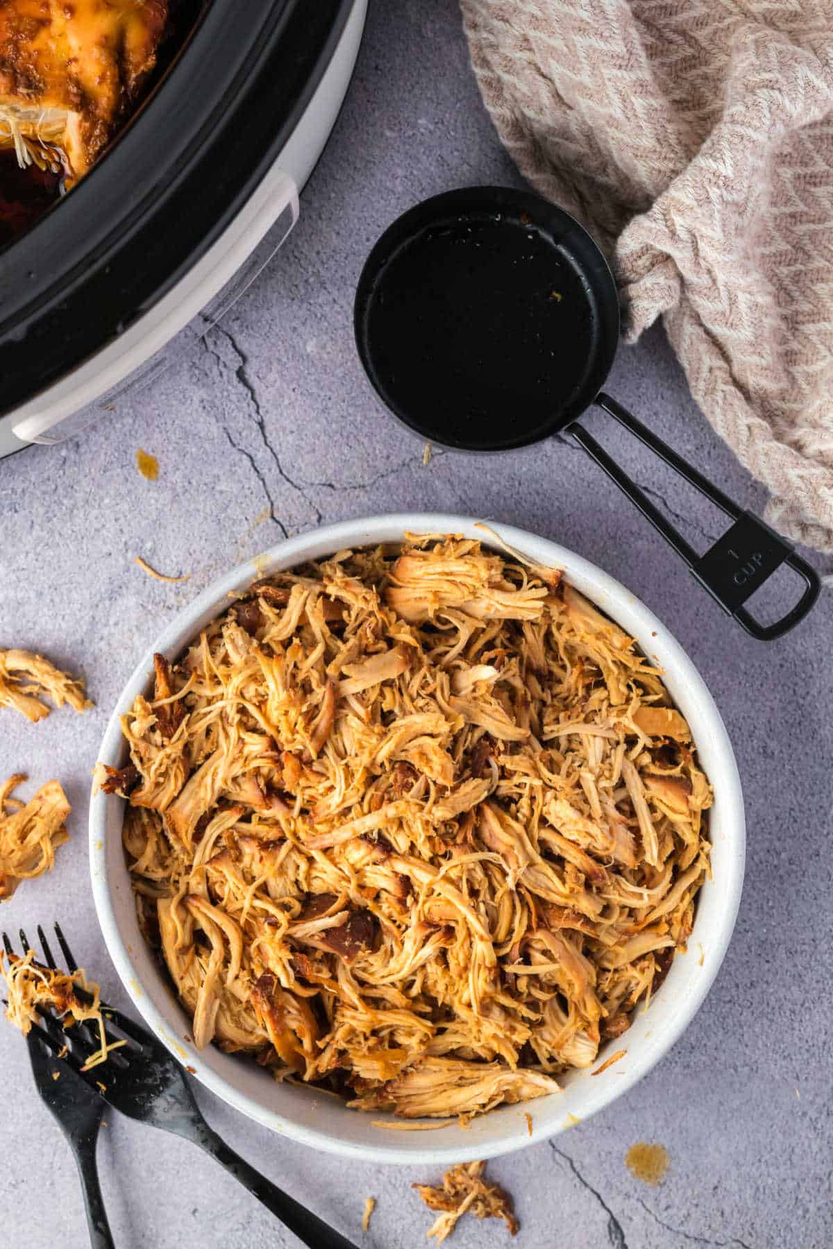 A bowl of shredded chicken, perfect for crockpot bbq chicken sliders, sits on a countertop next to a measuring cup and a cloth.