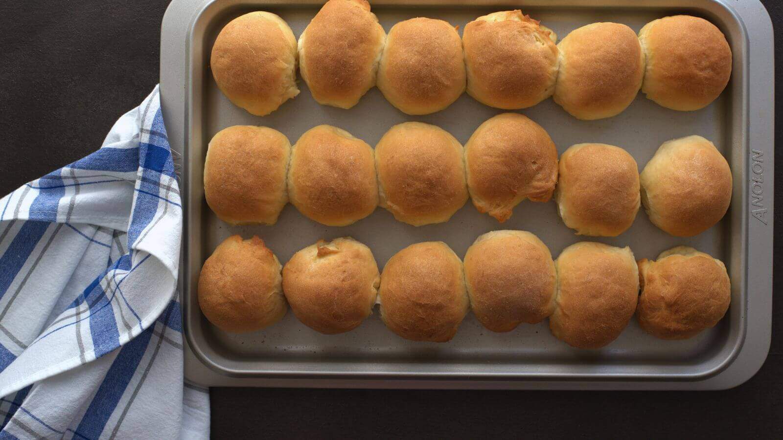 A baking tray with three rows of dinner rolls, next to a white and blue checkered cloth.