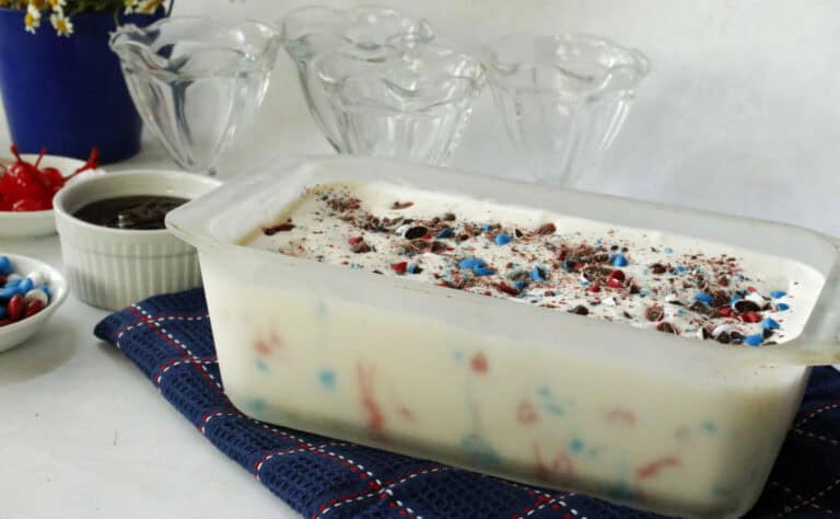 A container of vanilla ice cream with red and blue sprinkles on a blue cloth, with dessert bowls, cherries, and chocolate sauce in the background.