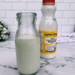 A glass bottle and a plastic bottle of Kalona Super Natural organic buttermilk are placed on a kitchen counter with a white tile backsplash.