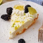 A slice of creamy pie garnished with whipped cream, lemon and lime slices, and blackberries is on a white speckled plate.