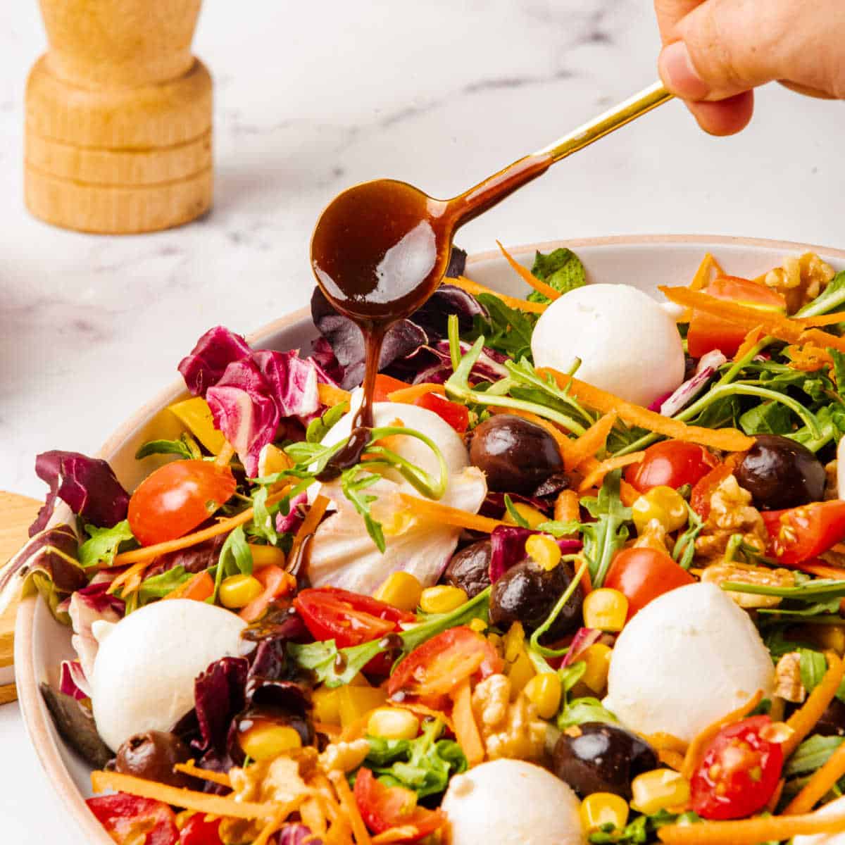 A hand drizzles balsamic dressing over a fresh salad containing lettuce, cherry tomatoes, corn, olives, shredded carrots, walnuts, and mozzarella balls, with a wooden pepper grinder in the background.