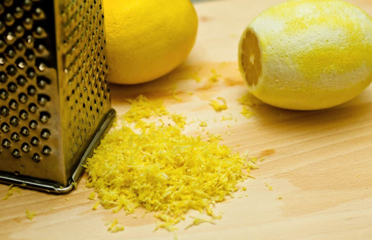 A metal grater, a pile of lemon zest, and two lemons, one partially zested, are placed on a wooden surface.