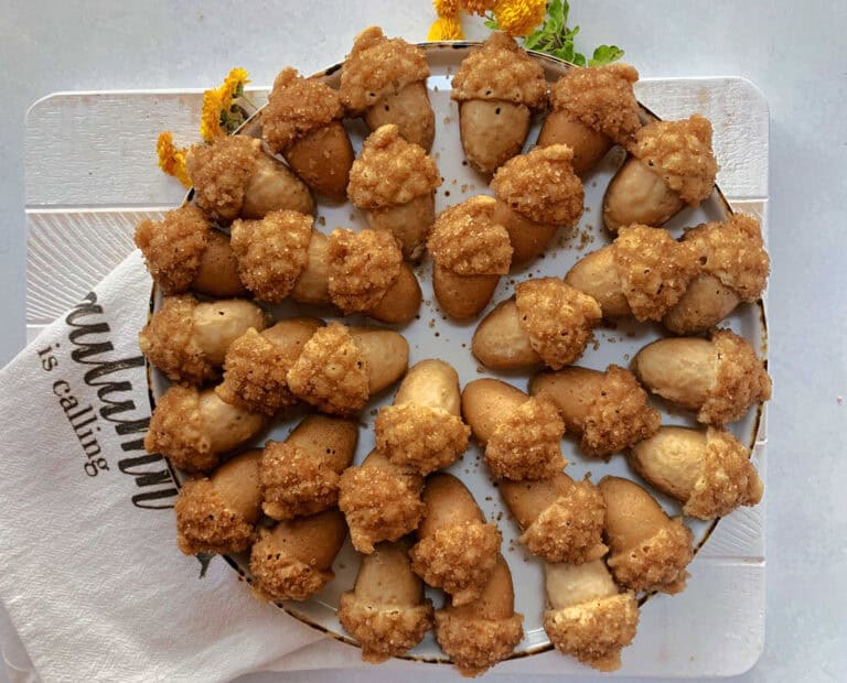 A plate filled with acorn-shaped cookies, each topped with a textured, crumbly coating, is surrounded by a white cloth napkin and yellow flowers.