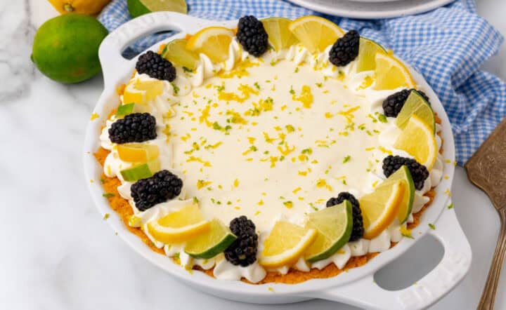 A key lime cheesecake garnished with lime slices, orange slices, blackberries, and whipped cream sits on a white plate with a blue checkered cloth underneath.