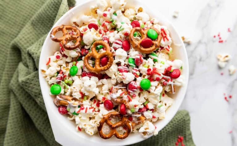 A bowl of popcorn mixed with red and green candies, pretzels, and red sprinkles, placed on a green cloth.