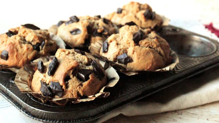 Five chocolate chunk muffins in paper liners sit in a muffin tin on a light-colored cloth.