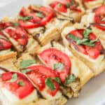 Slices of toasted bread topped with tomatoes, fresh basil, and melted cheese arranged on a white plate.