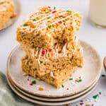 A stack of three square rice crispy treats with white icing and sprinkles on a beige ceramic plate. More treats and a glass of milk are in the background.