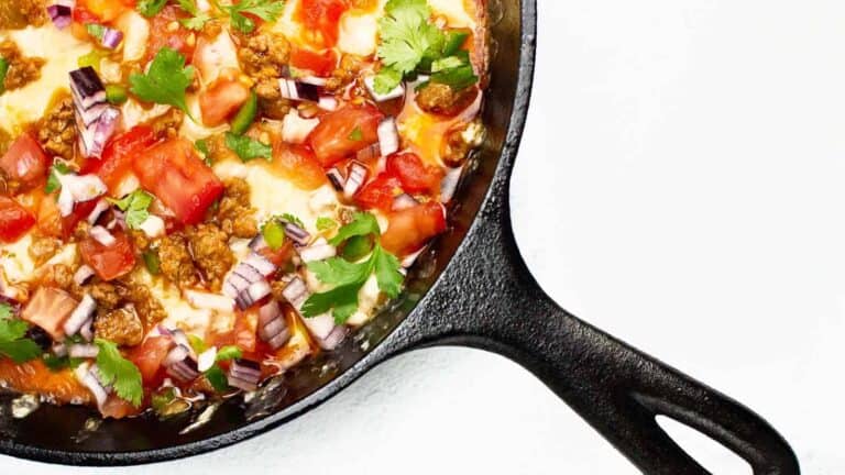 Skillet with melted cheese, chopped tomatoes, red onions, and cilantro on top of a white surface.