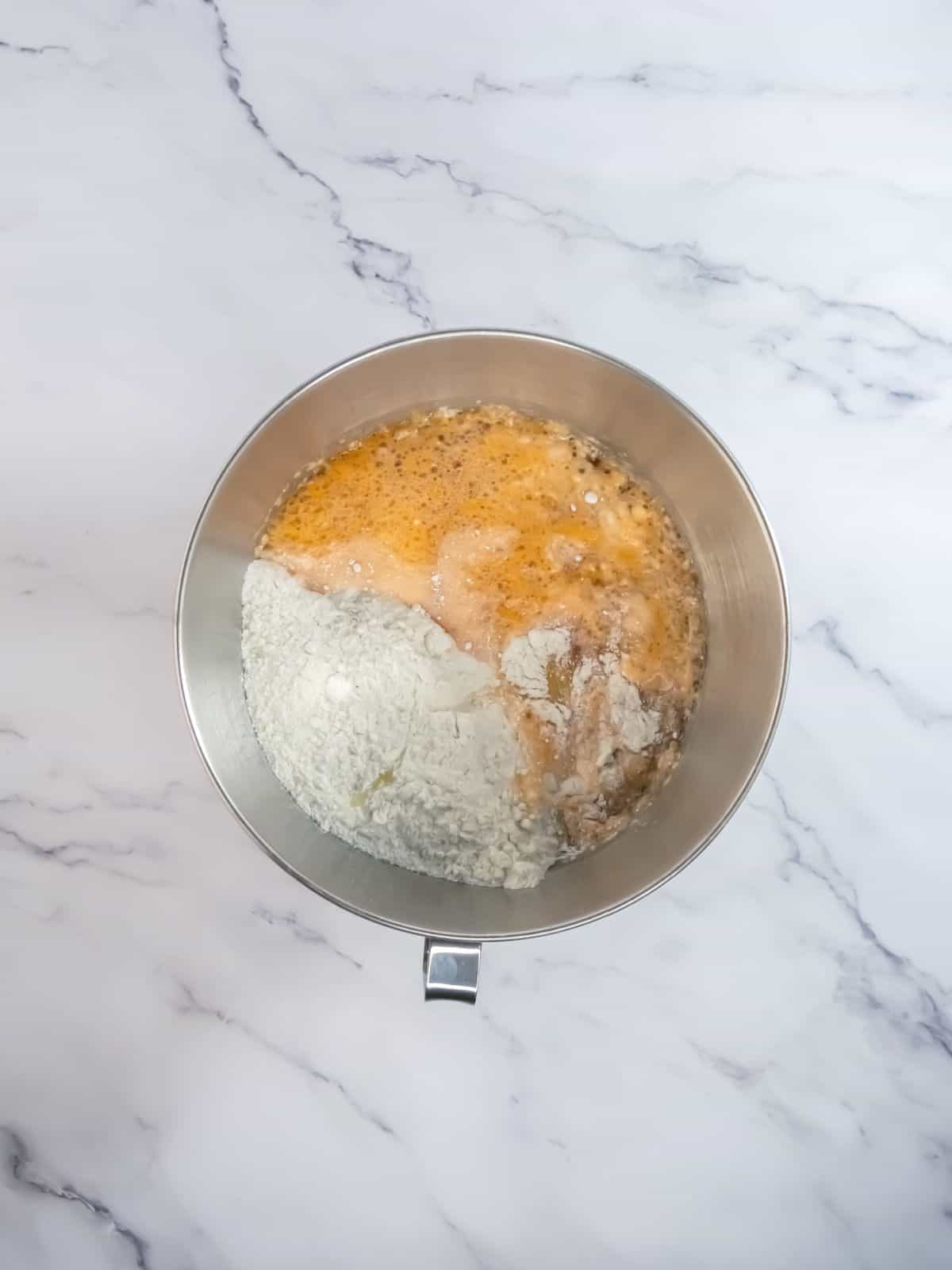 A mixing bowl containing flour, yeast, foamy liquid, and other ingredients placed on a marble countertop.