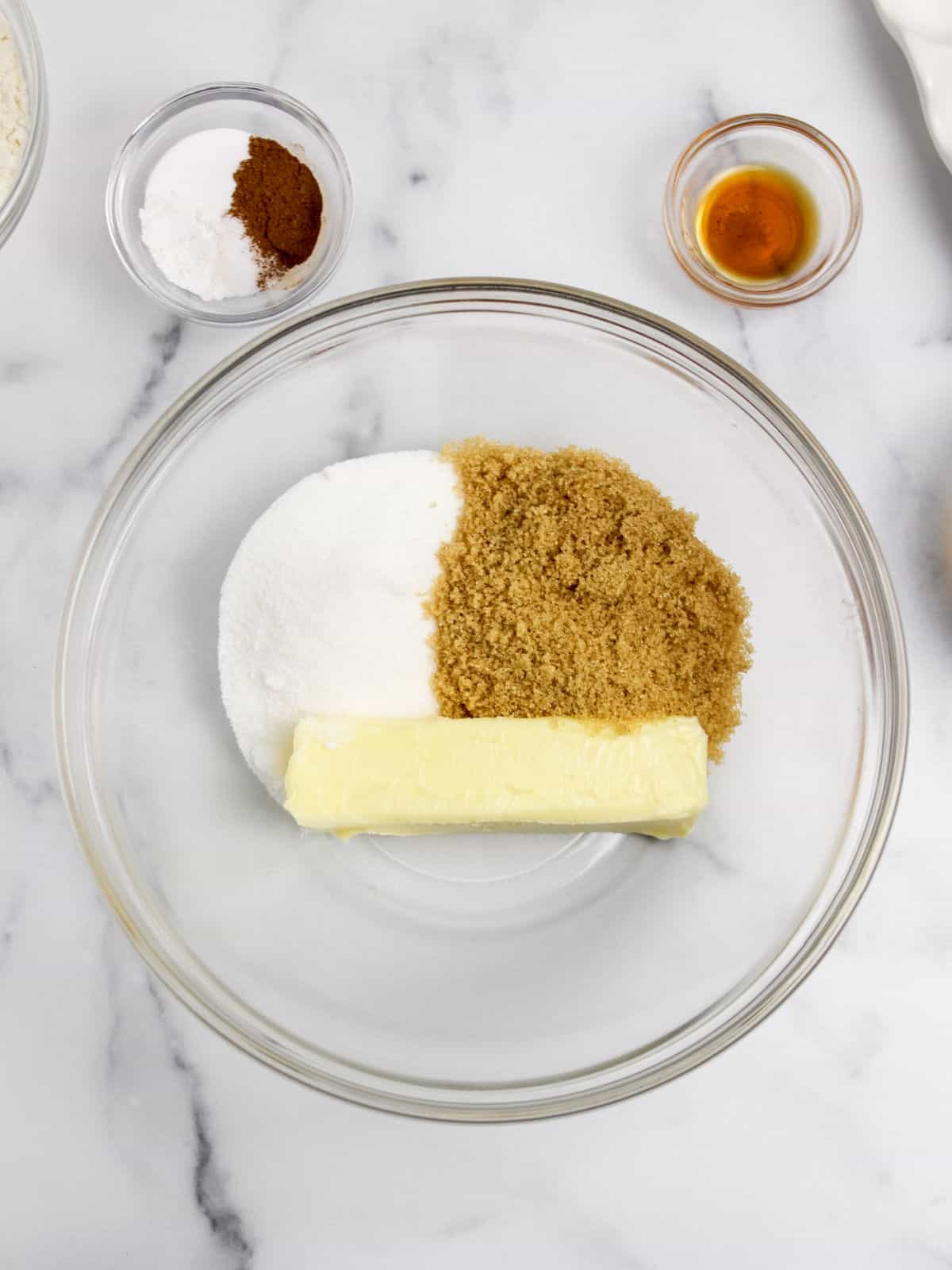 A glass bowl with sugar, brown sugar, and a stick of butter. Nearby, there are small bowls containing spices, baking powder, and vanilla extract, all arranged on a marble surface.