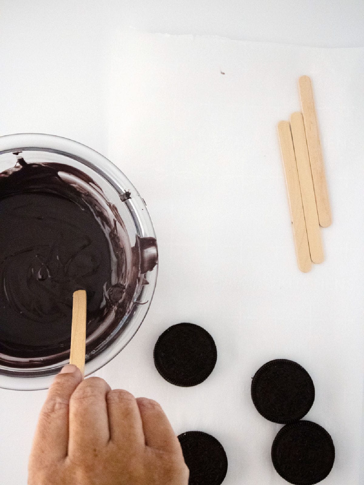 A hand dips a popsicle stick into melted chocolate in a bowl, crafting delightful Halloween Oreo cauldron pops. Four chocolate sandwich cookies and more popsicle sticks lie on a white surface nearby.