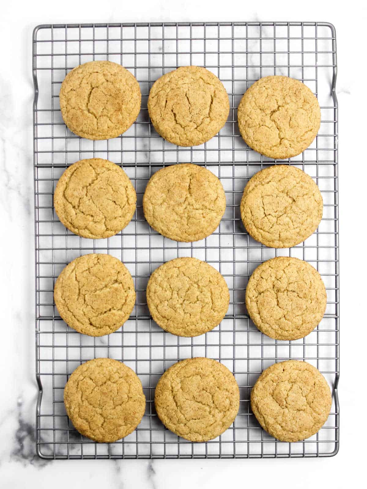 A cooling rack with twelve evenly spaced, freshly baked Snickderdoodle cookies on a white marble surface.