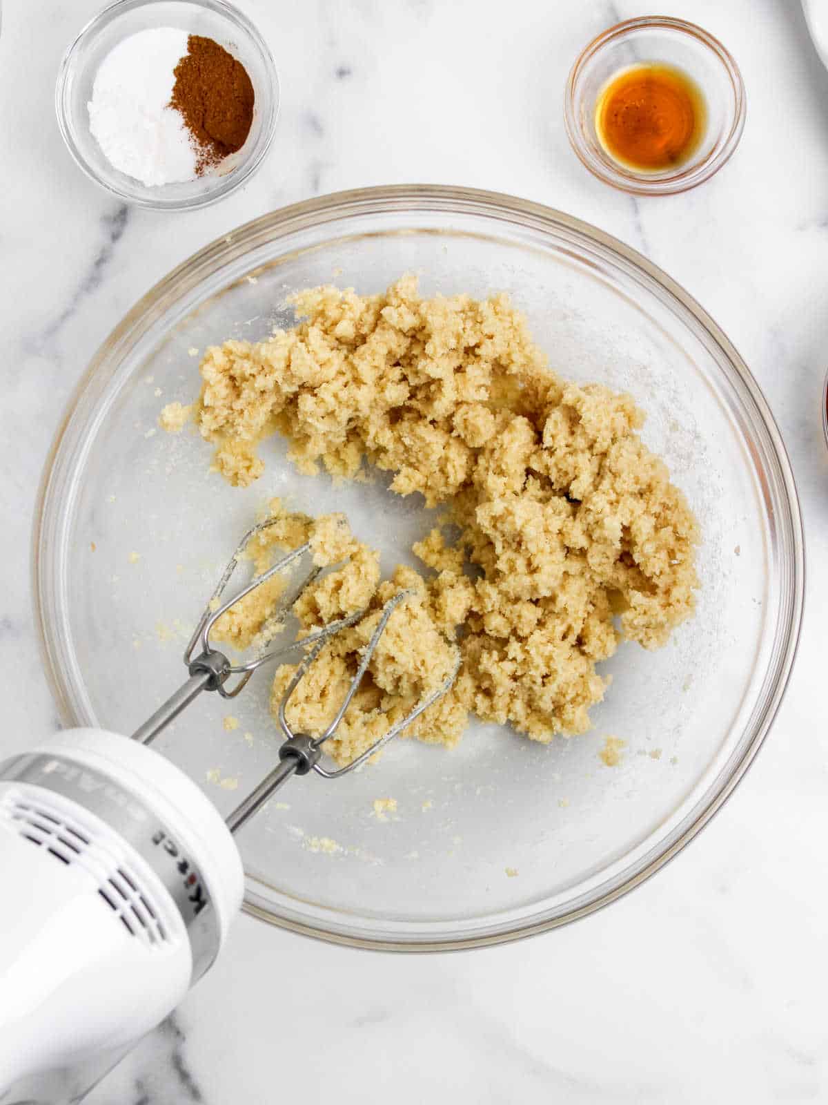 An electric hand mixer in a glass bowl with a crumbly mixture of ingredients on a marble countertop, surrounded by small bowls containing vanilla extract, baking powder, and cinnamon.