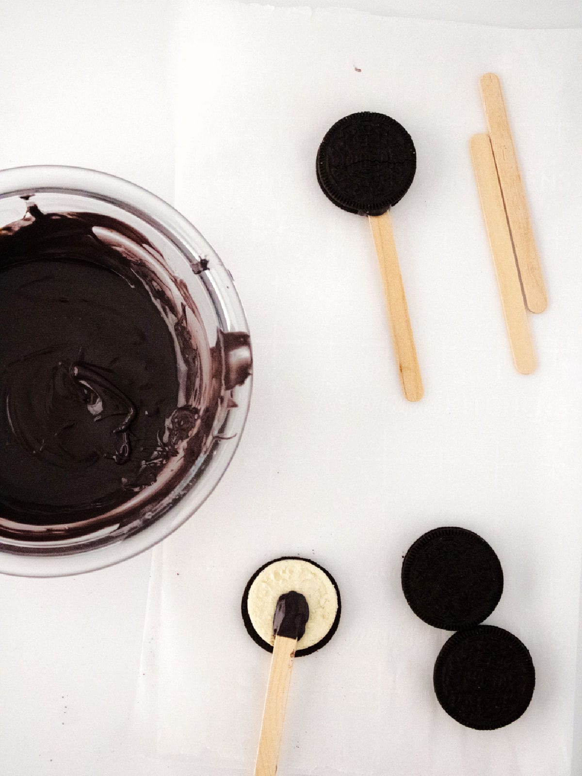 A bowl of melted chocolate next to three Halloween cauldron pops being made with chocolate sandwich cookies and wooden sticks on a white surface.