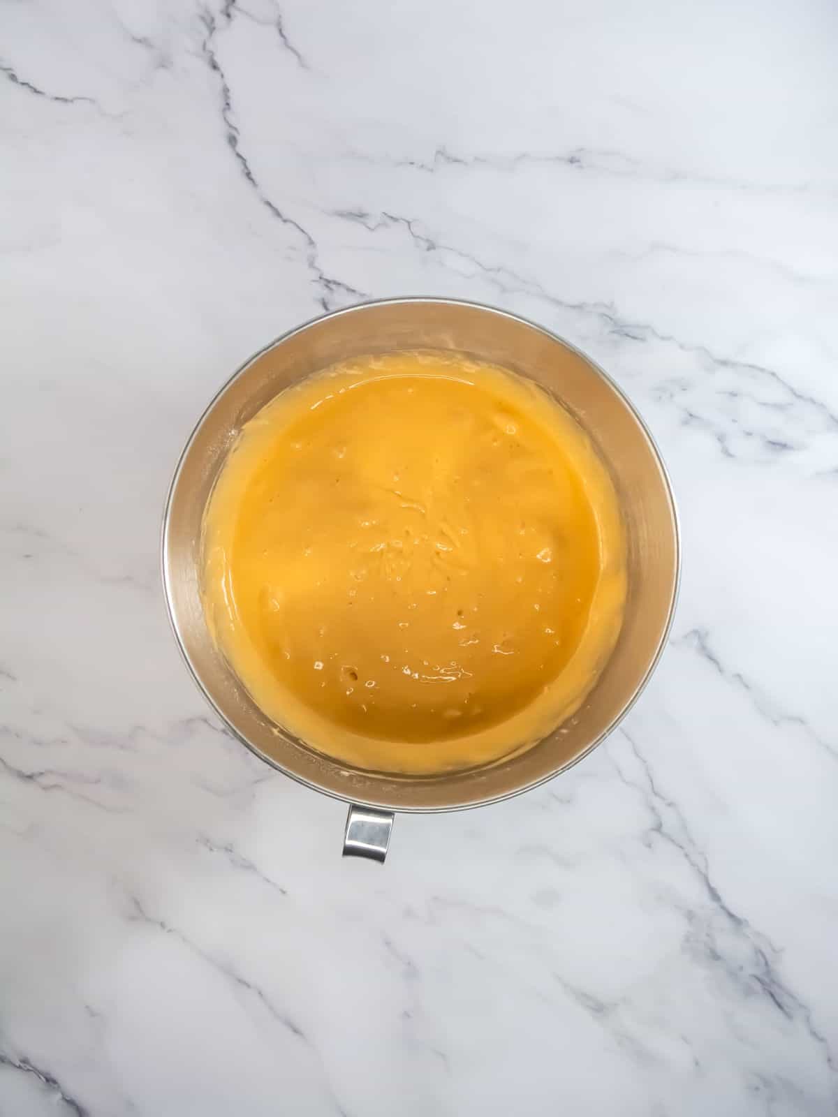 A stainless steel mixing bowl containing well-mixed yellow cake batter sits on a marble countertop.