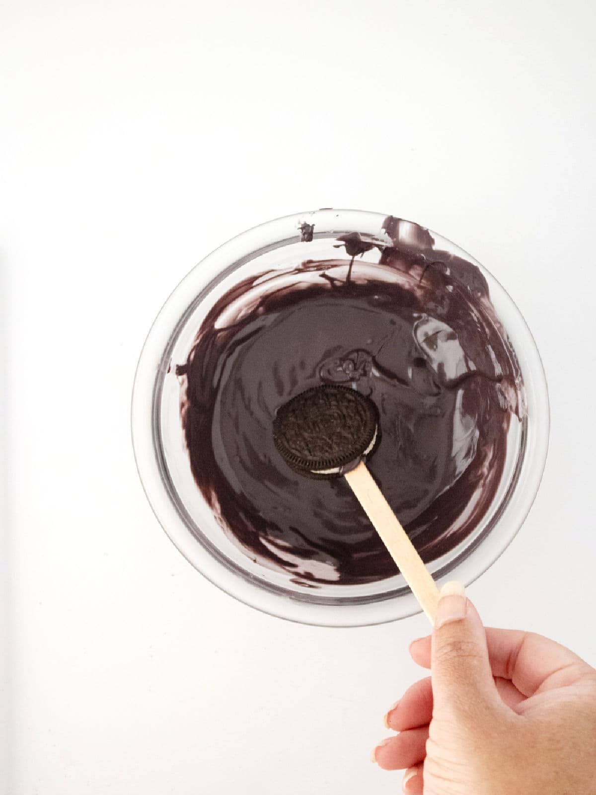 A hand holds a wooden stick with an Oreo cookie dipped in melted chocolate in a glass bowl, conjuring Halloween cauldron pops against a white background.