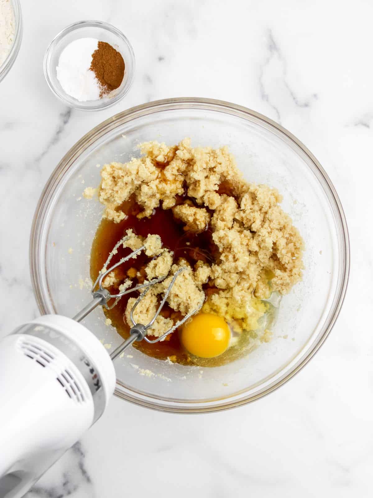 A glass mixing bowl with raw cookie dough, an egg, and a hand mixer. Nearby, a small bowl contains cinnamon and baking powder on a marble countertop.