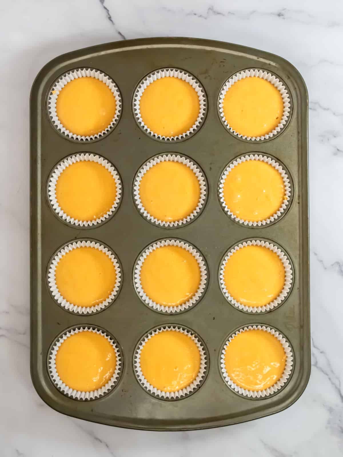 A 12-cup muffin tin filled with yellow batter in white paper liners, placed on a marble surface.