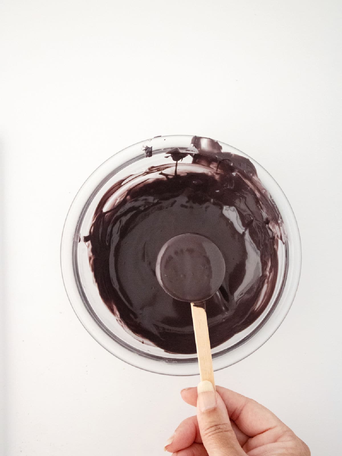 A hand holding a wooden spoon over a bowl of dark liquid, possibly melted chocolate, on a white surface—perfect for making Halloween cauldron pops.
