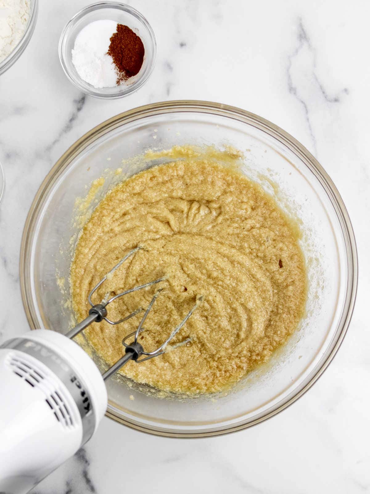 A bowl of beige batter is being mixed with an electric hand mixer. A small glass dish containing cocoa powder and baking soda is placed nearby on a marble countertop.