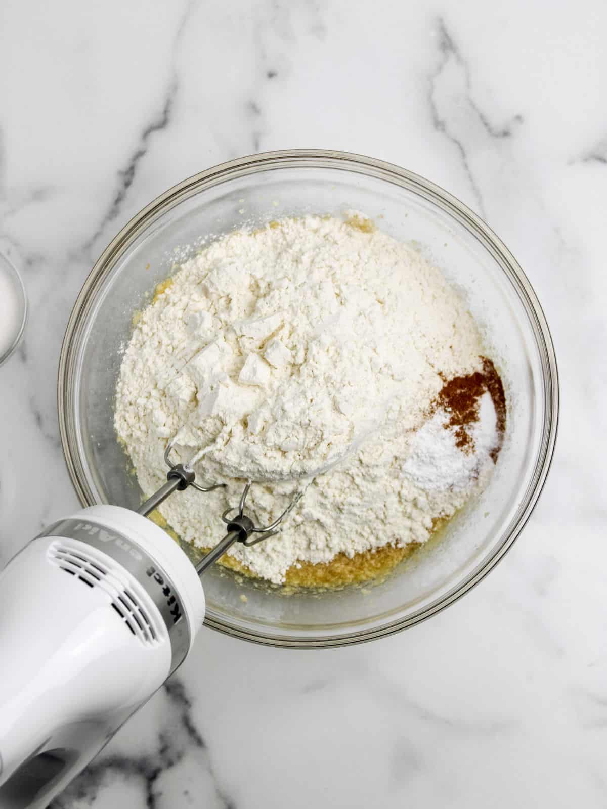 A glass bowl containing flour, spices, and other ingredients is being mixed with an electric hand mixer on a marble countertop.