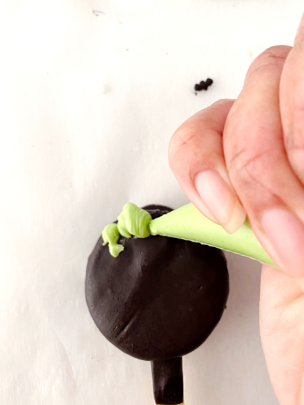 A hand holding a green piping bag decorates a round black Halloween Oreo on a white surface.