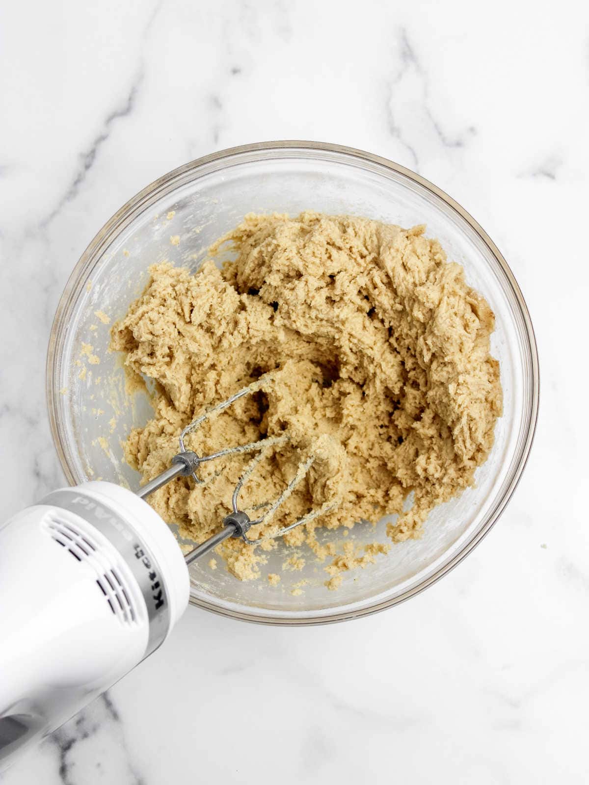 A glass mixing bowl filled with partially mixed cookie dough being blended with an electric hand mixer on a white marble surface.
