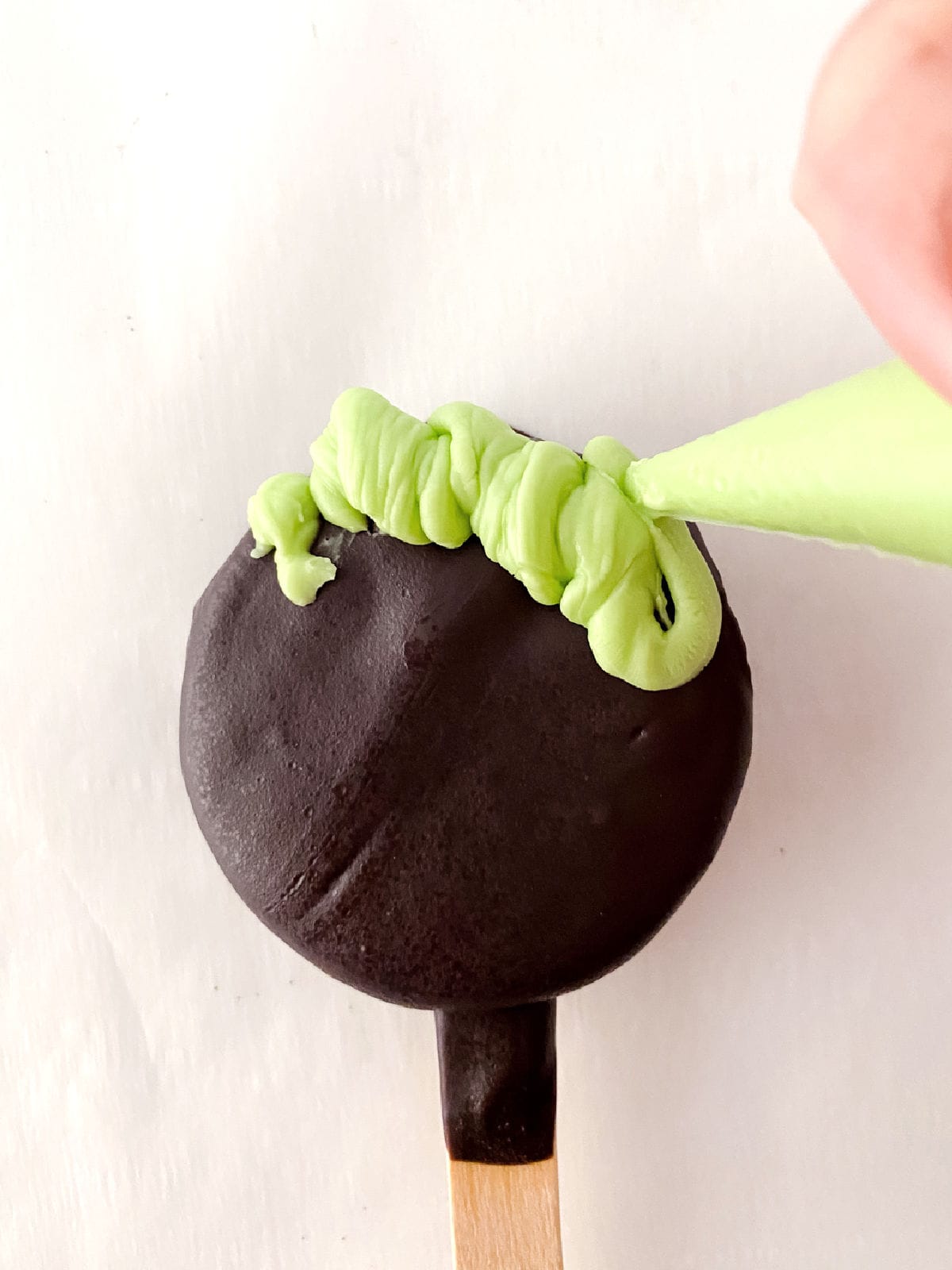 A hand piping green frosting onto a round, chocolate-coated treat on a wooden stick, transforming it into a Halloween cauldron pop.