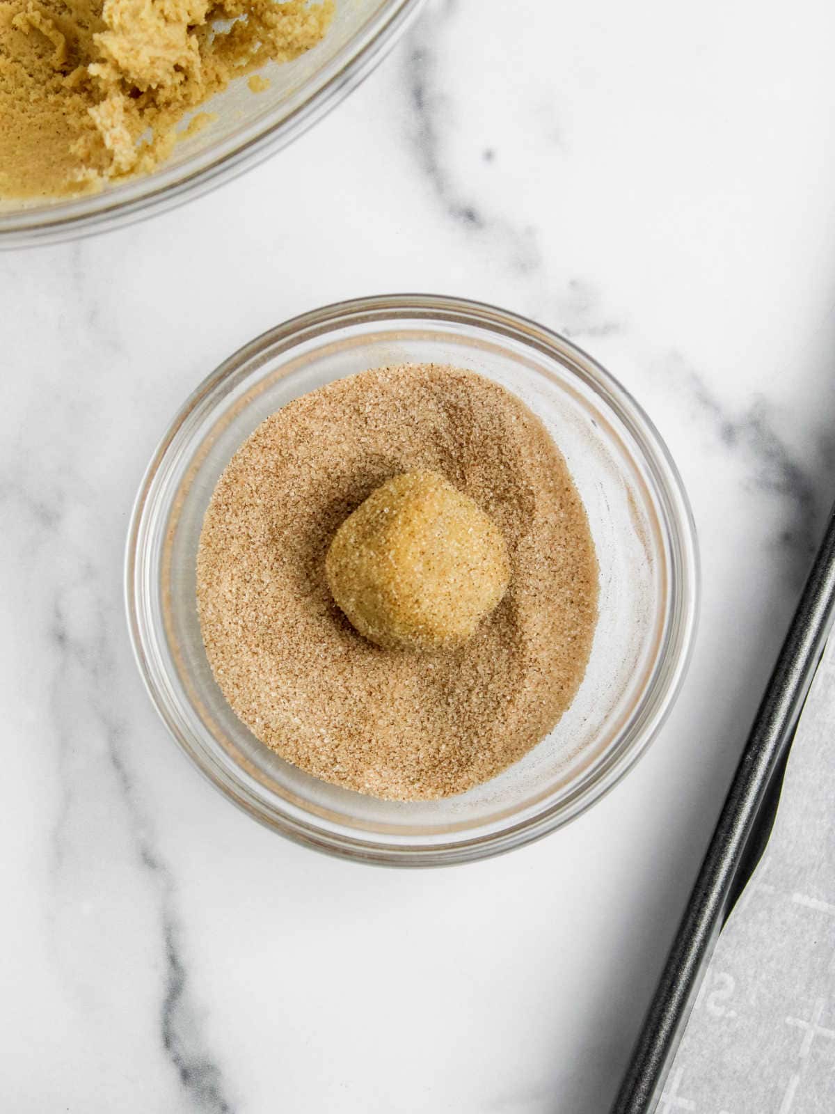 A cookie dough ball being rolled in a bowl of cinnamon sugar.