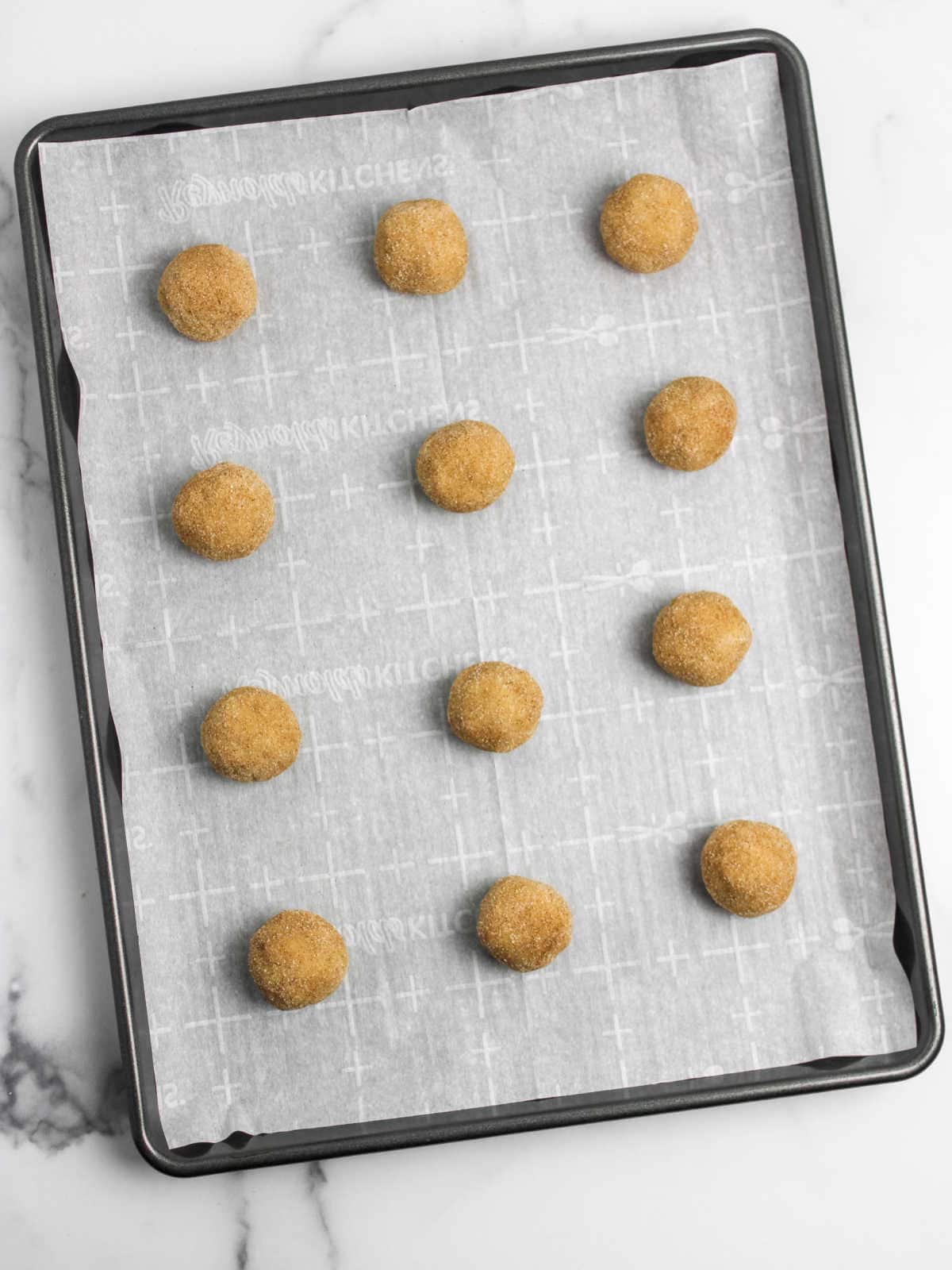A baking sheet lined with parchment paper holds 12 evenly spaced, round dough balls covered in a sugar-cinnamon coating.